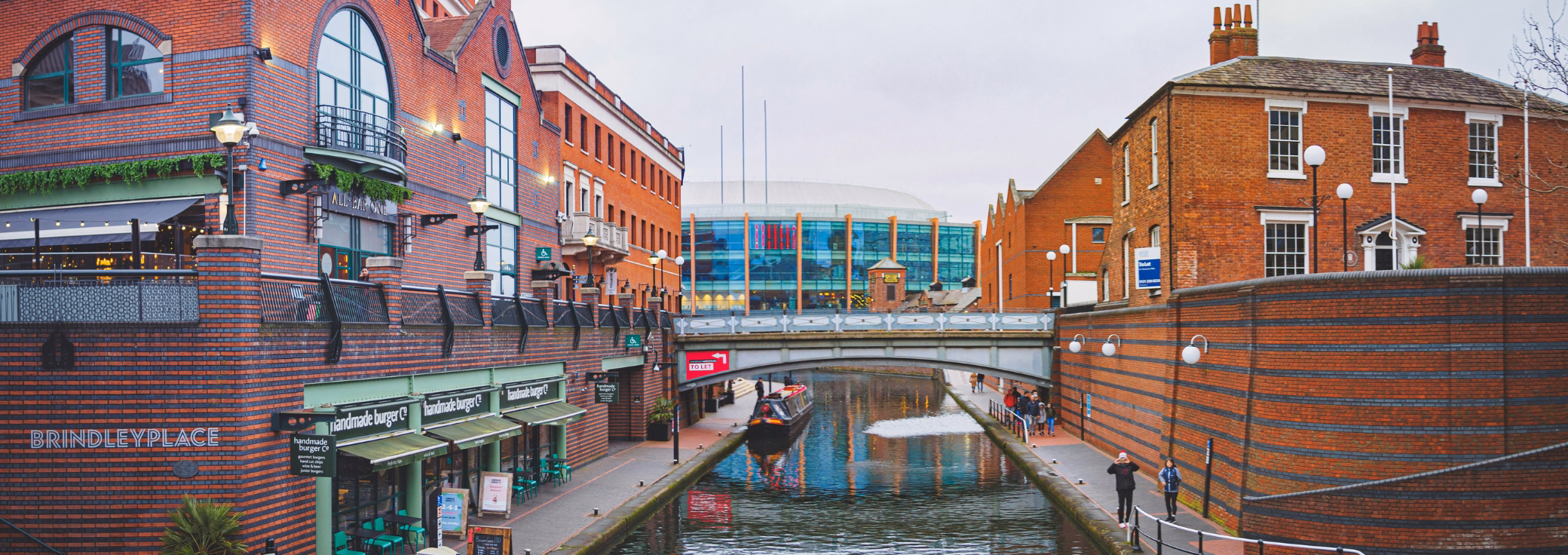 People walk on paths beside a canal with a boat under a bridge. Brick buildings stand on both sides, and green chairs sit outside a restaurant.
