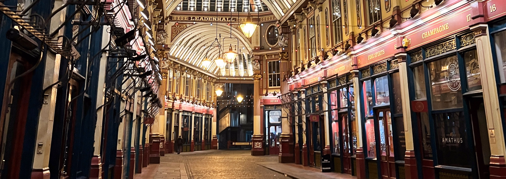 A person walks past red and blue storefronts on a street under a roof with lights and text reading LEADENHALL.