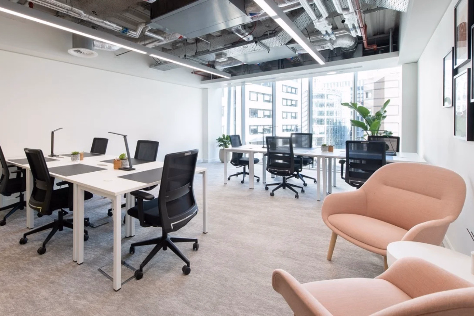 Black chairs at white desks and pink armchairs on a grey floor near floor-to-ceiling windows under pipes and lights.