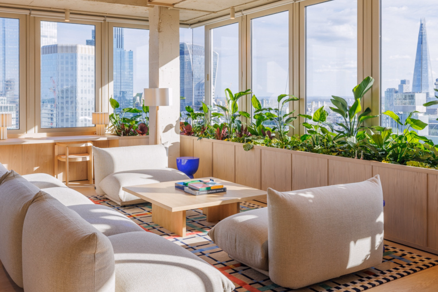 Cream sofas and a wood table sit on a rug near windows. Plants near a view of skyscrapers (The Shard).