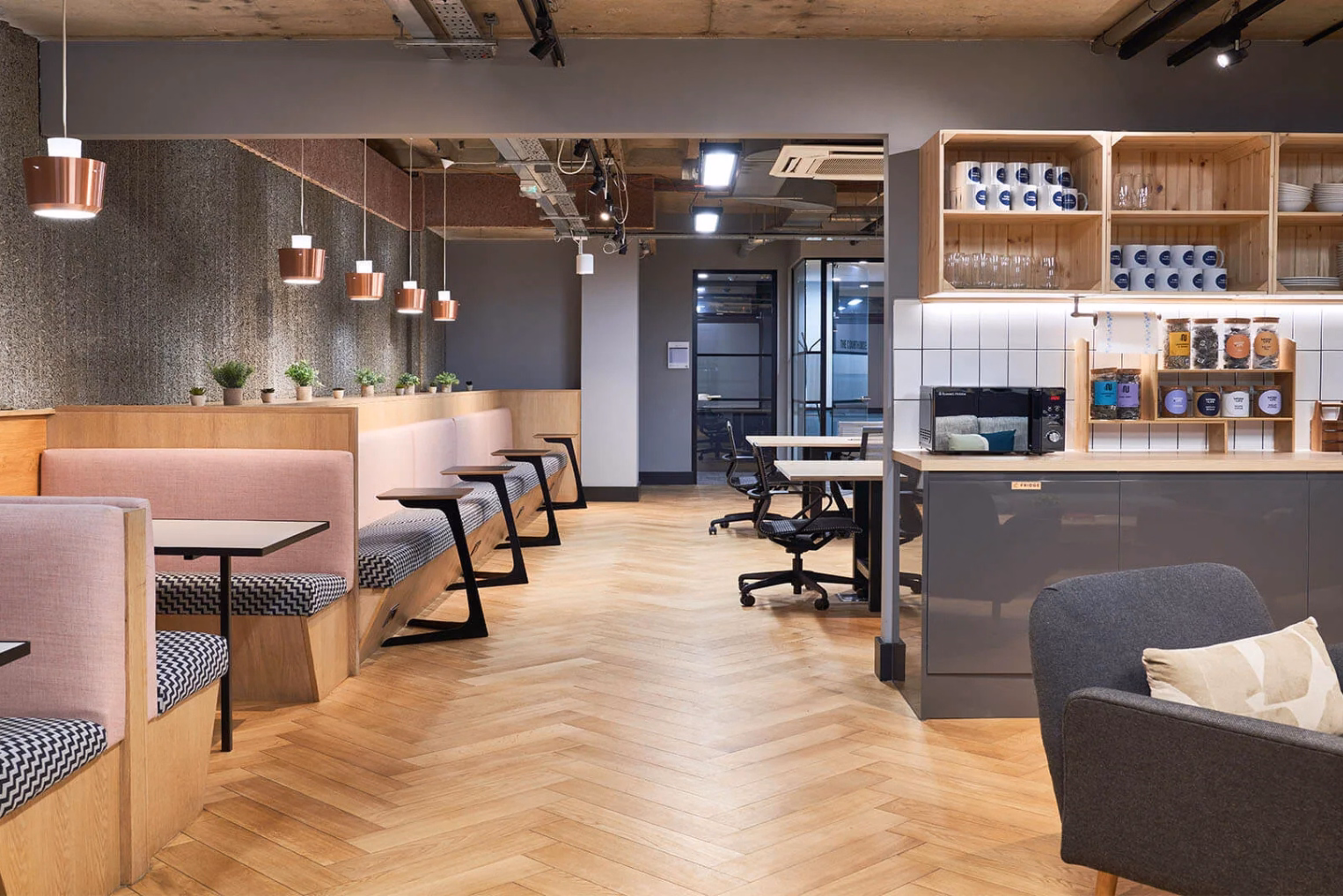 Pink booths and black tables sit on a wood floor. A grey armchair, a grey counter, and black chairs stand under lights.