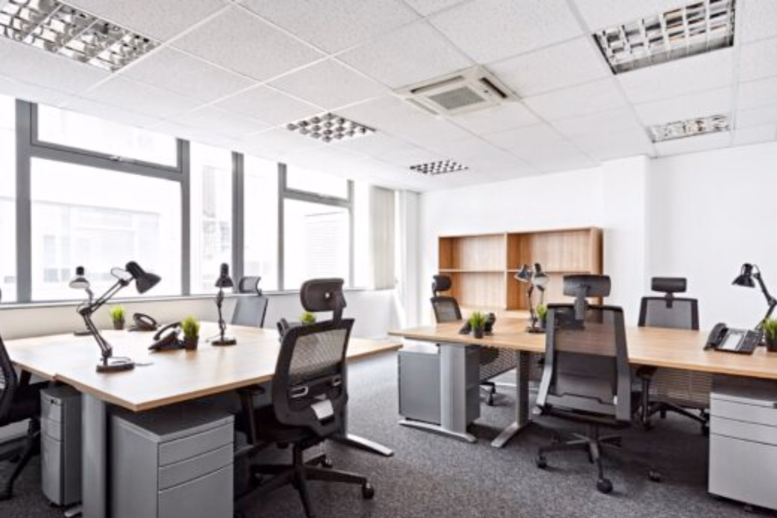 Black chairs sit at wooden desks on a grey floor. Grey cabinets and a wood bookshelf stand near windows under ceiling lights.