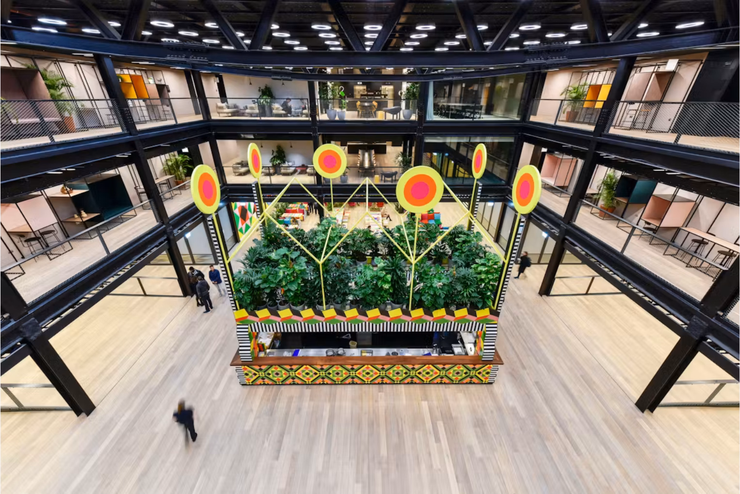 People walk on a wood floor around an atrium with plants. Pink, yellow, and green booths stand on balconies under ceiling lights.