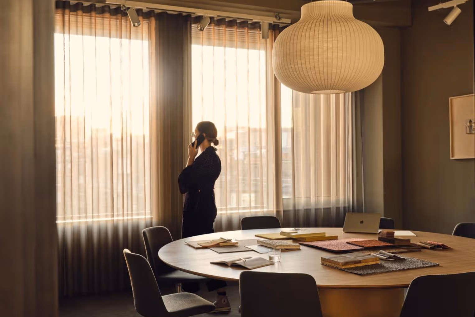 A woman talks on a phone near windows. Grey chairs sit at a wooden table under a white light.