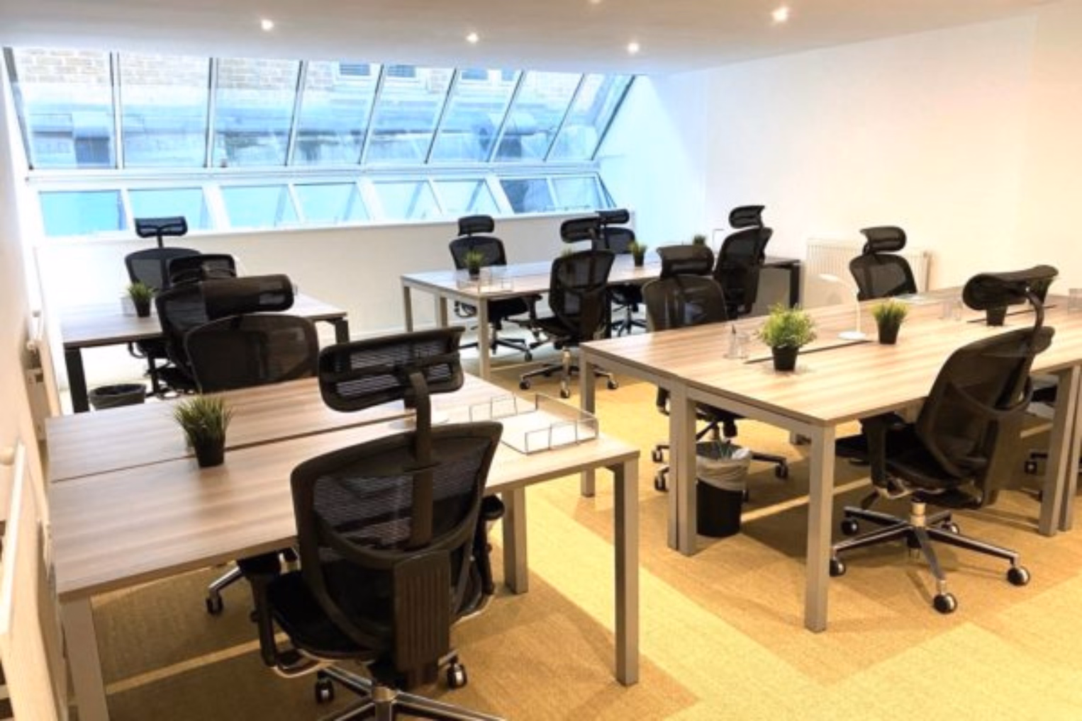 Black chairs at brown desks on a tan floor near a slanted window wall under ceiling lights.