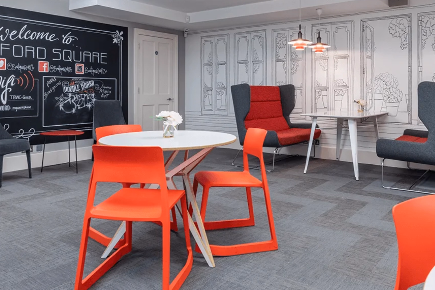 Orange chairs at a white table on a grey floor. Red and grey armchairs near a white table and a blackboard wall.