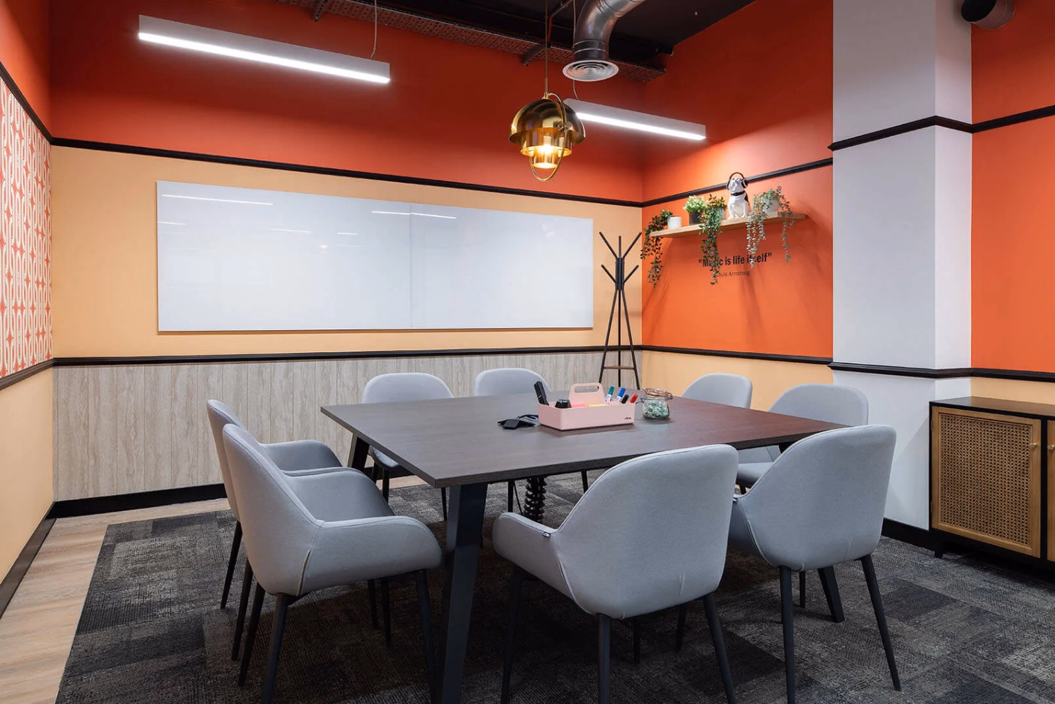 Grey chairs sit at a brown table on a grey carpet. A whiteboard hangs on an orange wall near a wooden cabinet.