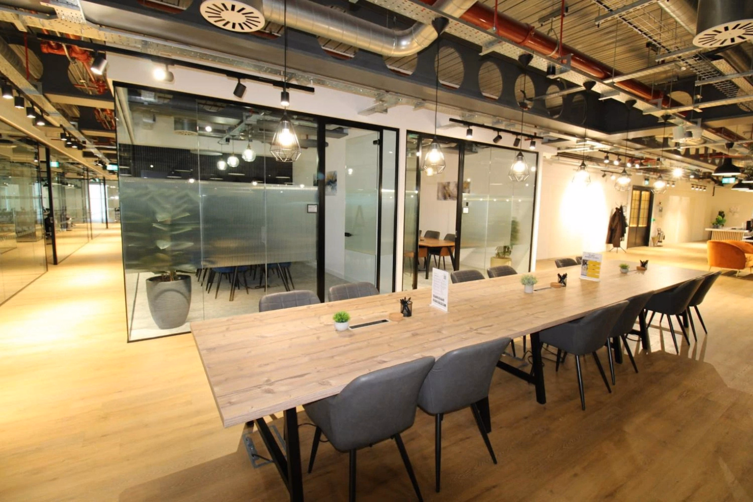 Grey chairs sit at a wooden table on a wood floor. Glass rooms and an orange armchair stand under ceiling pipes and lights.