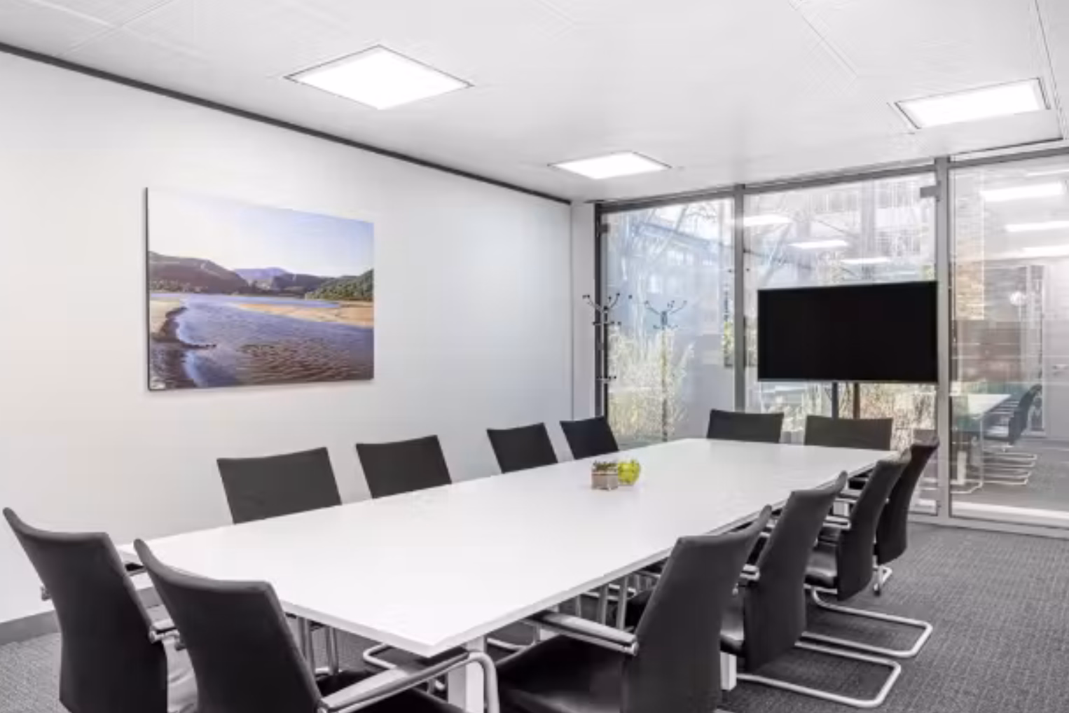Black chairs at a white table on a grey floor. A picture hangs on a wall near a black screen leaning against a glass wall.
