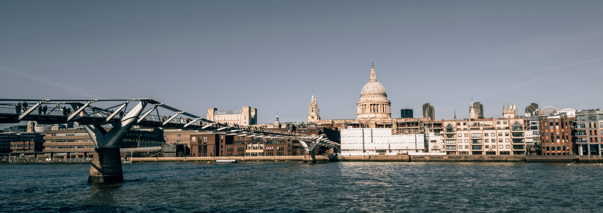 People walk on a bridge over a river toward a cathedral dome and buildings under a grey sky.