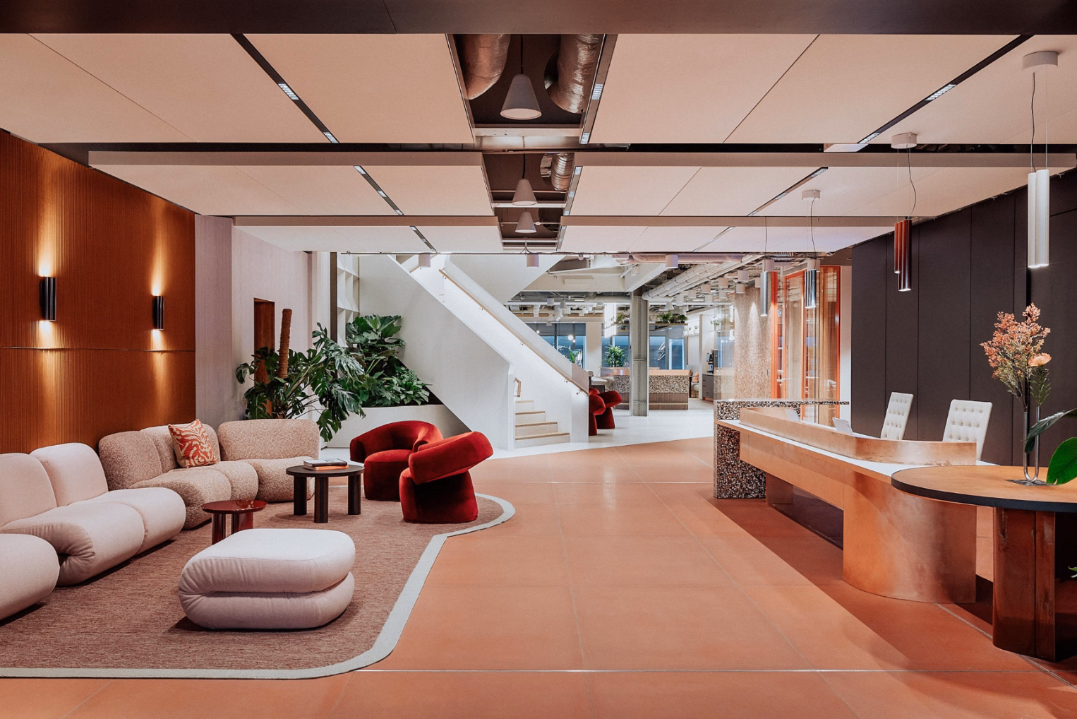 Pink and beige sofas, red armchairs, and dark wooden tables on a rug. A wood counter and white chairs stand on a tile floor near a staircase.