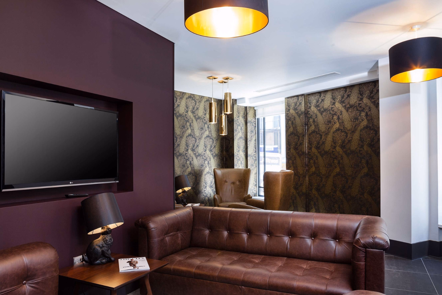 A burgundy sofa in the middle of the room beside a dark wood table with a black lamp on it. 2 tan armchairs stood under gold hanging lights.