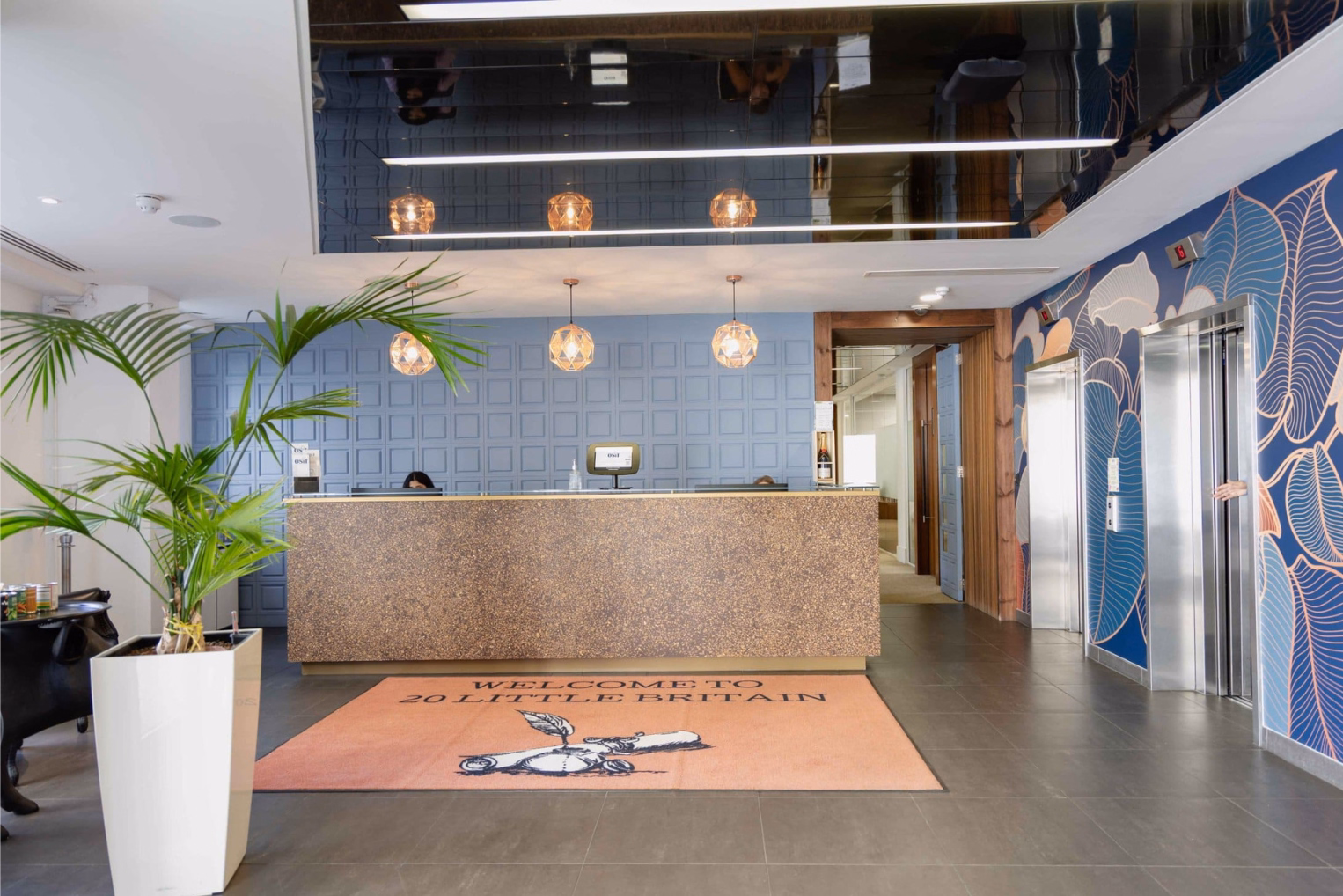 People sit behind a brown counter on a grey floor near an orange mat, a plant, and elevators under hanging lights.