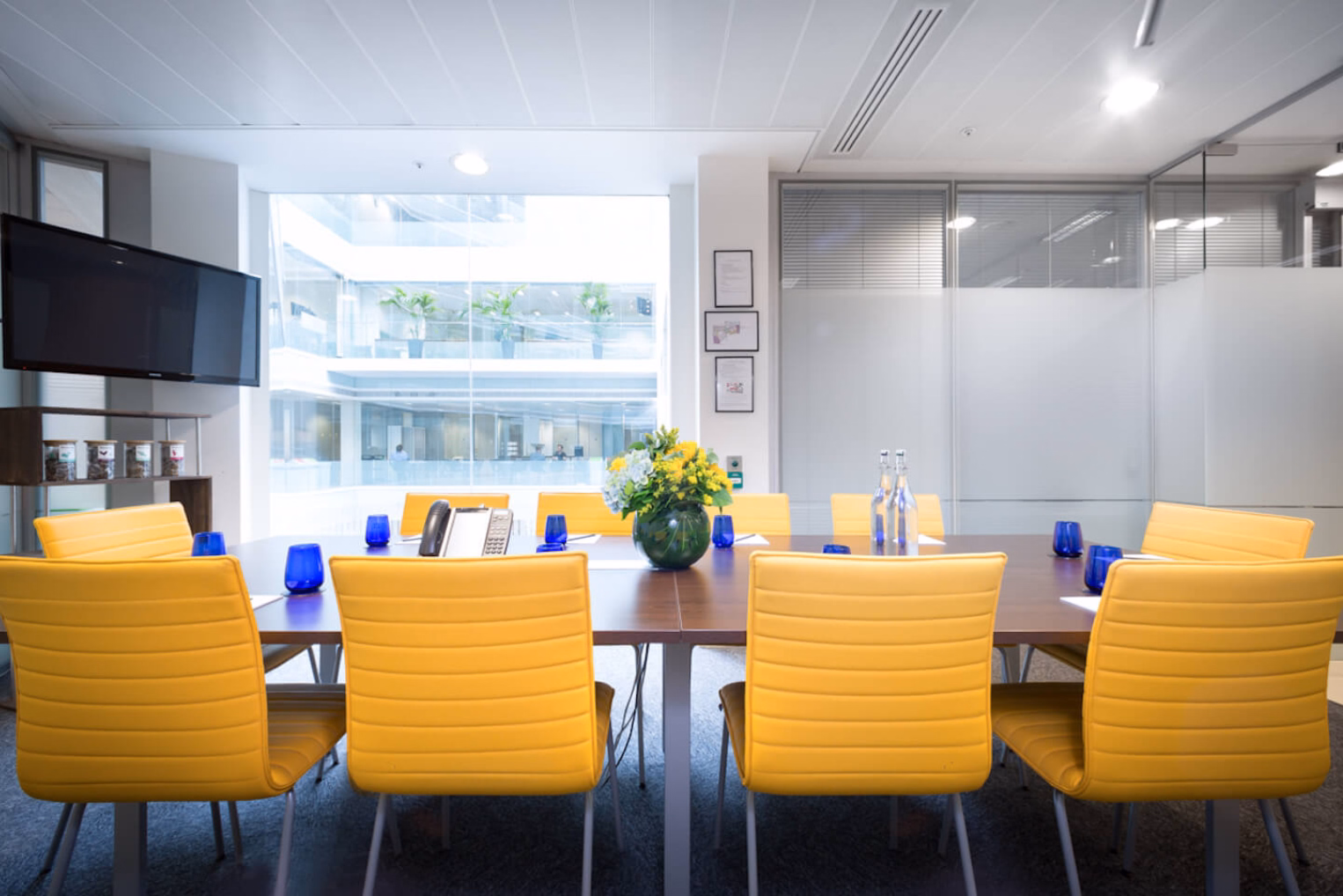 Yellow chairs sit at a brown table on a grey floor near a black screen and windows under lights.