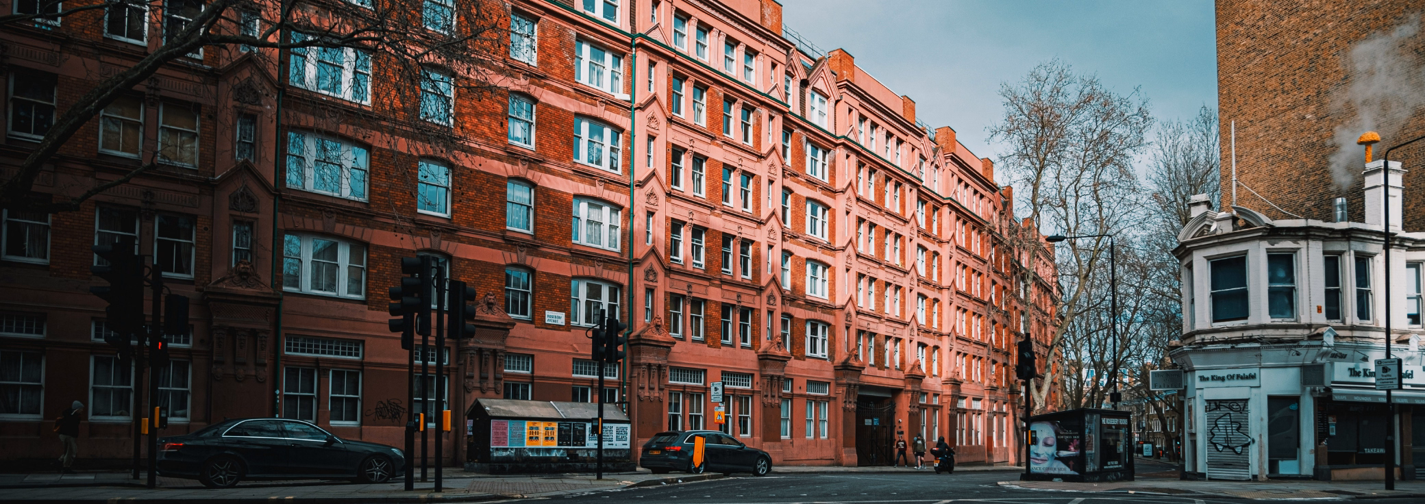 A car is parked, and people walk on a street near a red brick building and a white shop under a grey sky.