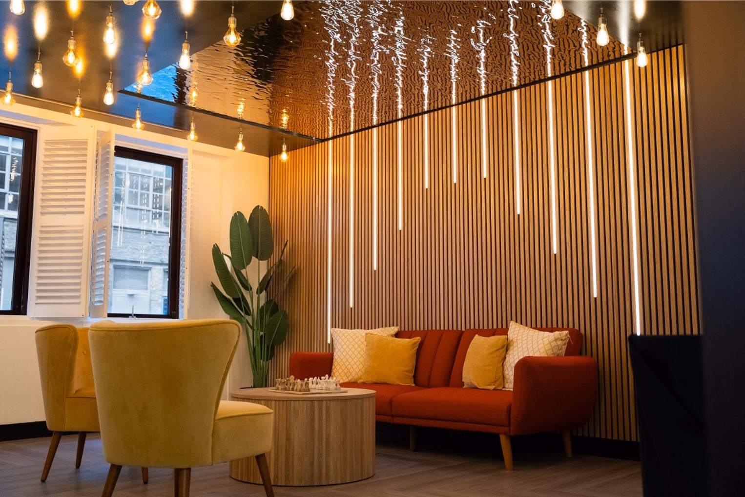 Yellow armchairs and a red sofa sit at a wooden table on the floor. A plant stands near a wooden wall under lights.