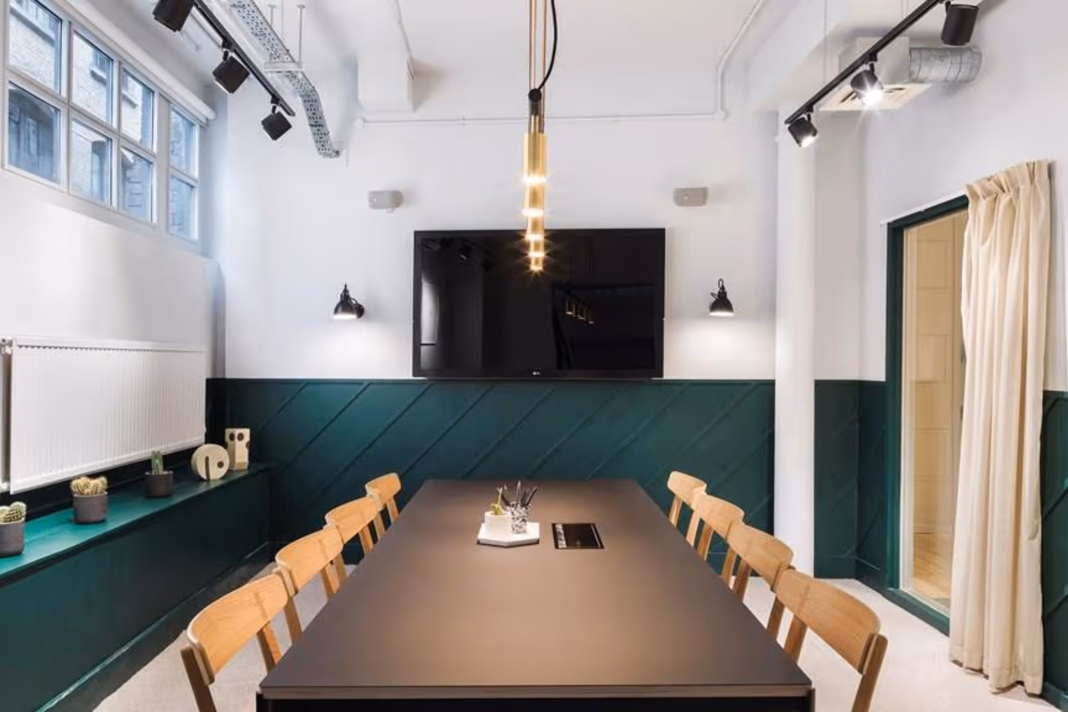 Wooden chairs at a black table on a white floor near a black screen and a green wall under lights.