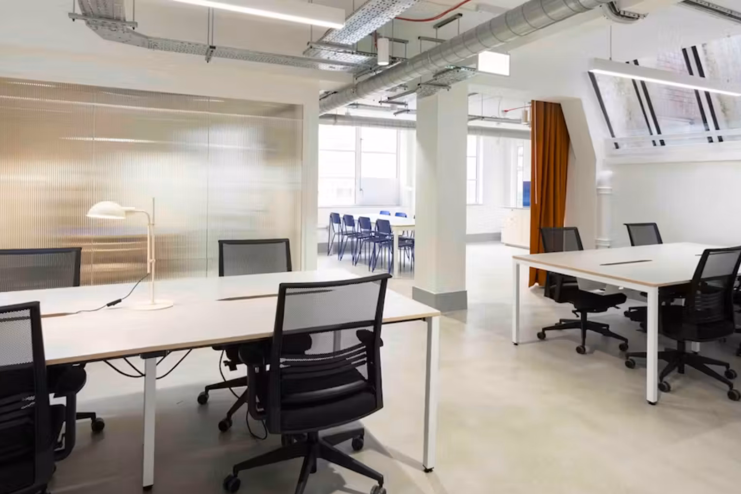 Black chairs at white desks on a grey floor. Blue chairs and a white table stand near an orange curtain under pipes and lights.