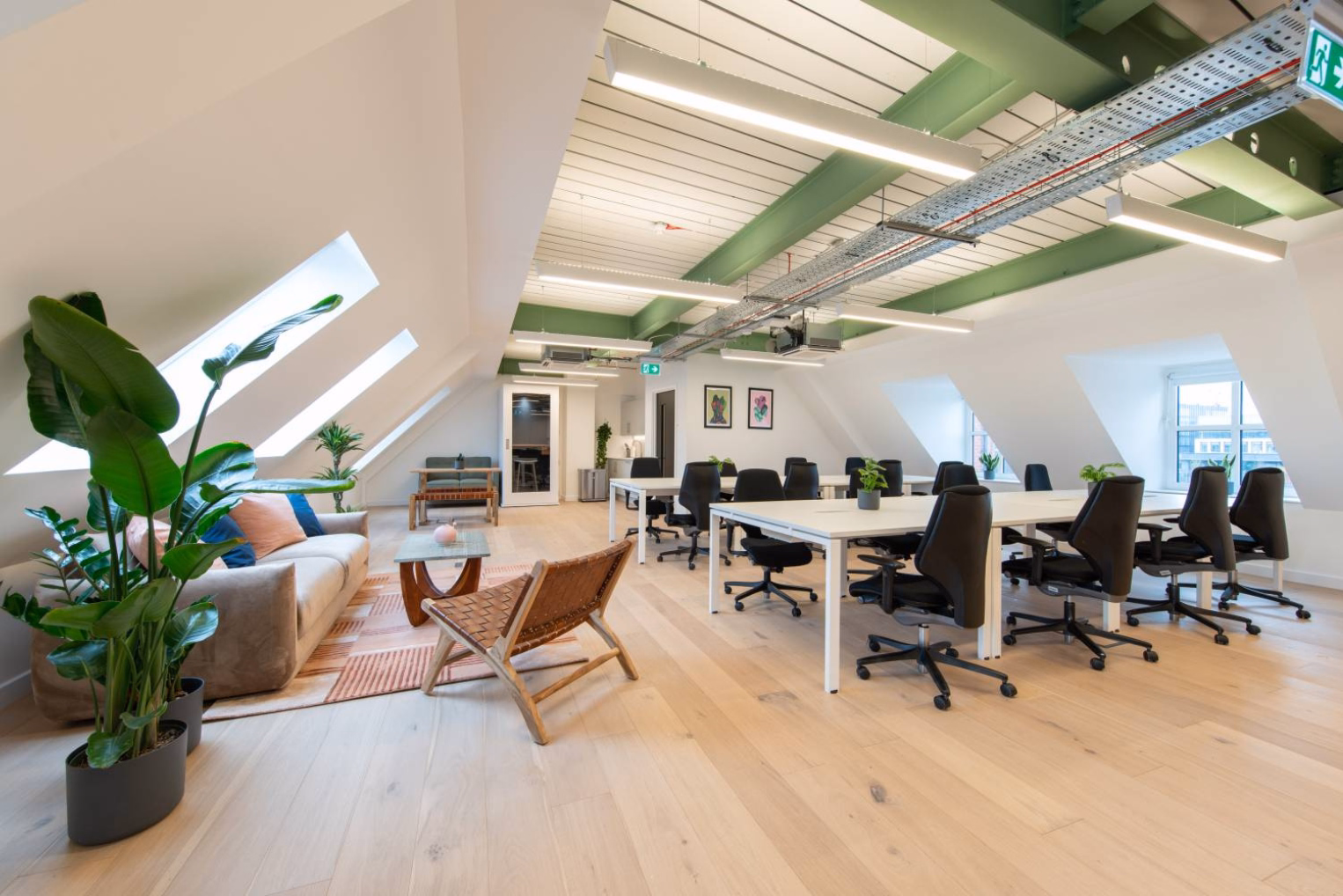 Black chairs at white desks on a wood floor. A beige sofa and a brown chair stand near windows under green beams and lights.