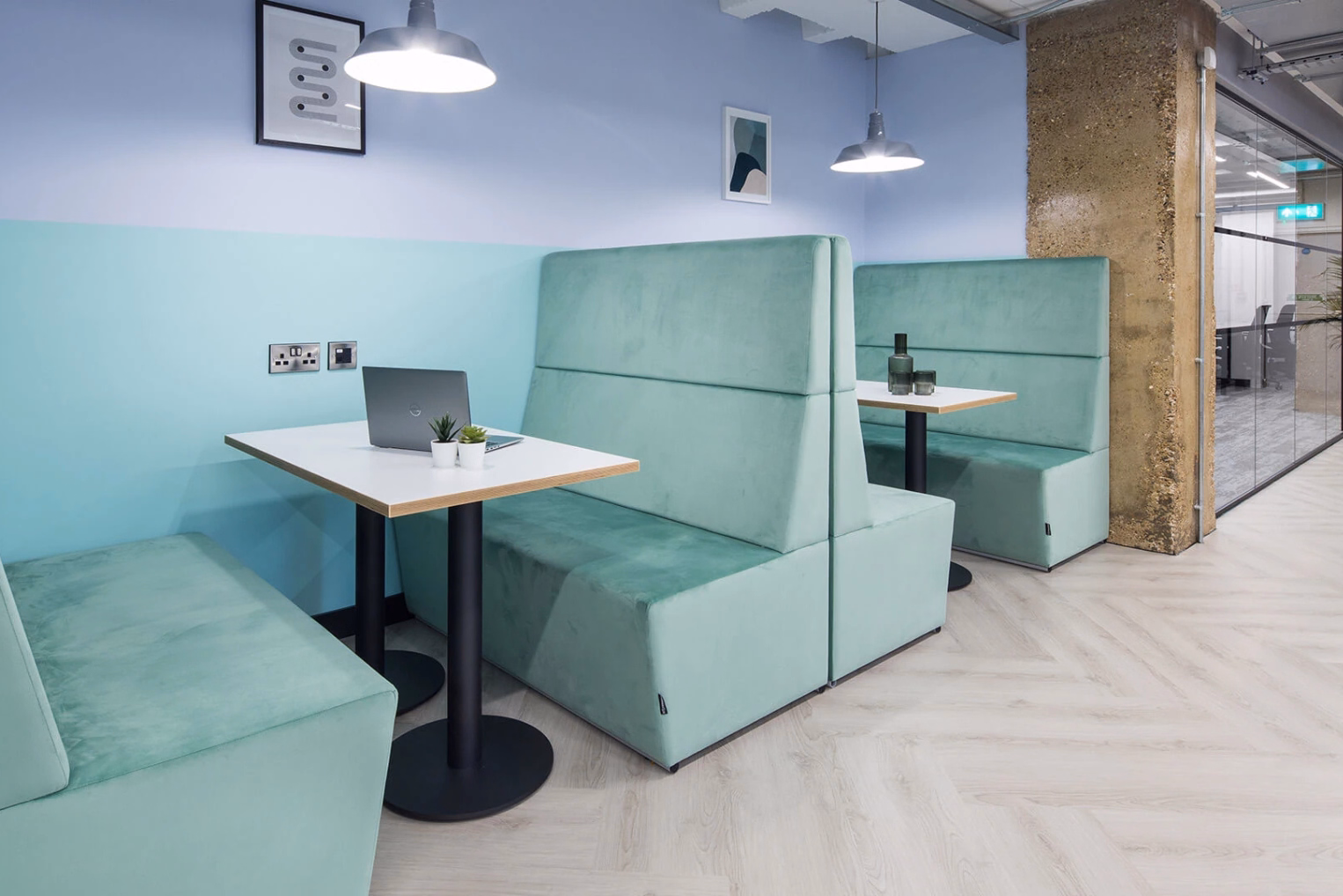 Teal blue booth sofas and white tables with a laptop, plants and a bottle on them, under hanging lights.