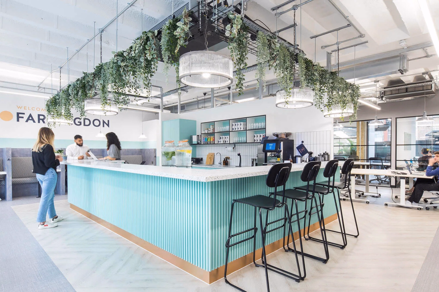 People talk at a green counter with black stools while a man sits at a white desk under plants and lights.