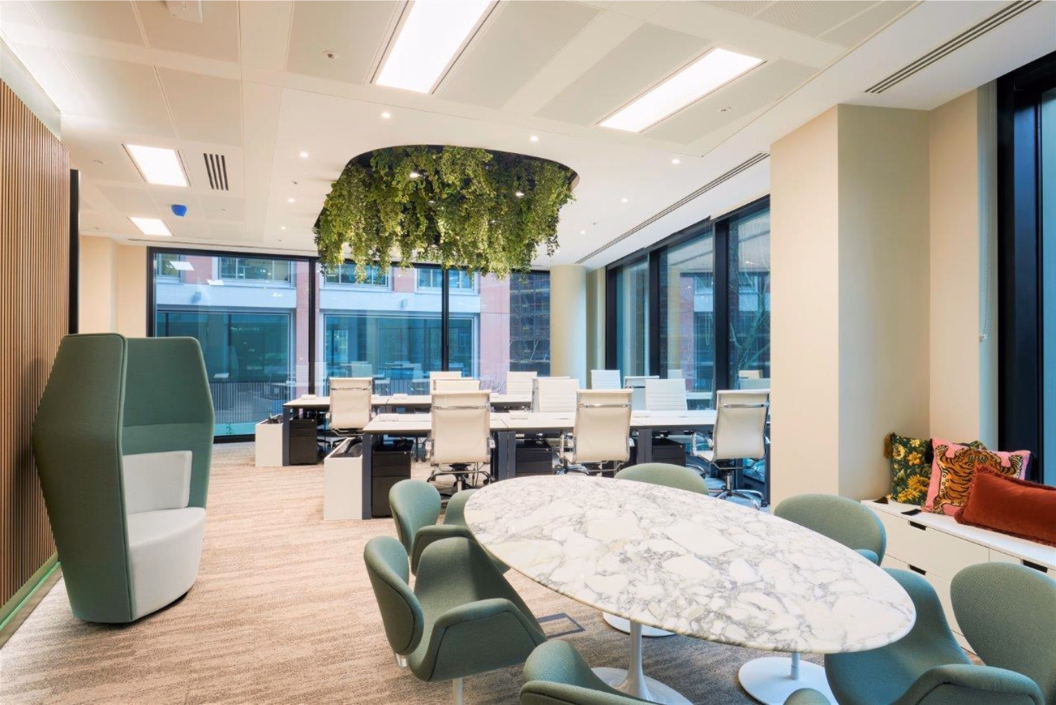 Green chairs sit at a white marble table. White chairs stand at white desks near windows, with a green pod beneath ceiling plants.