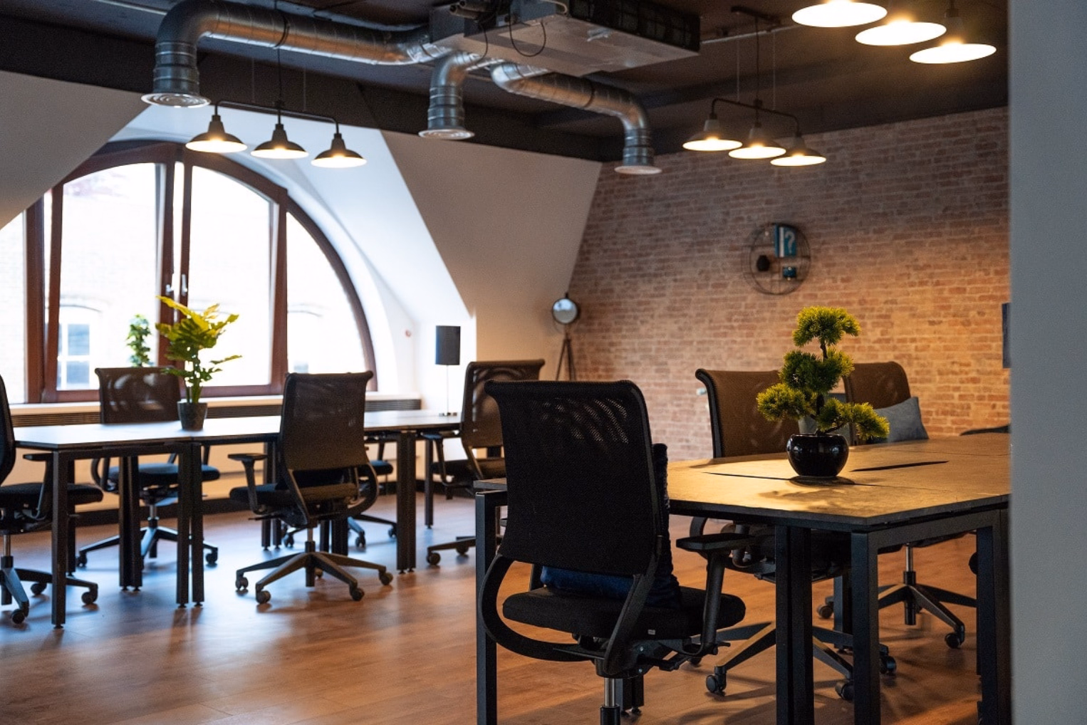 Black chairs at wooden desks below hanging ceiling lights and silver pipes.
