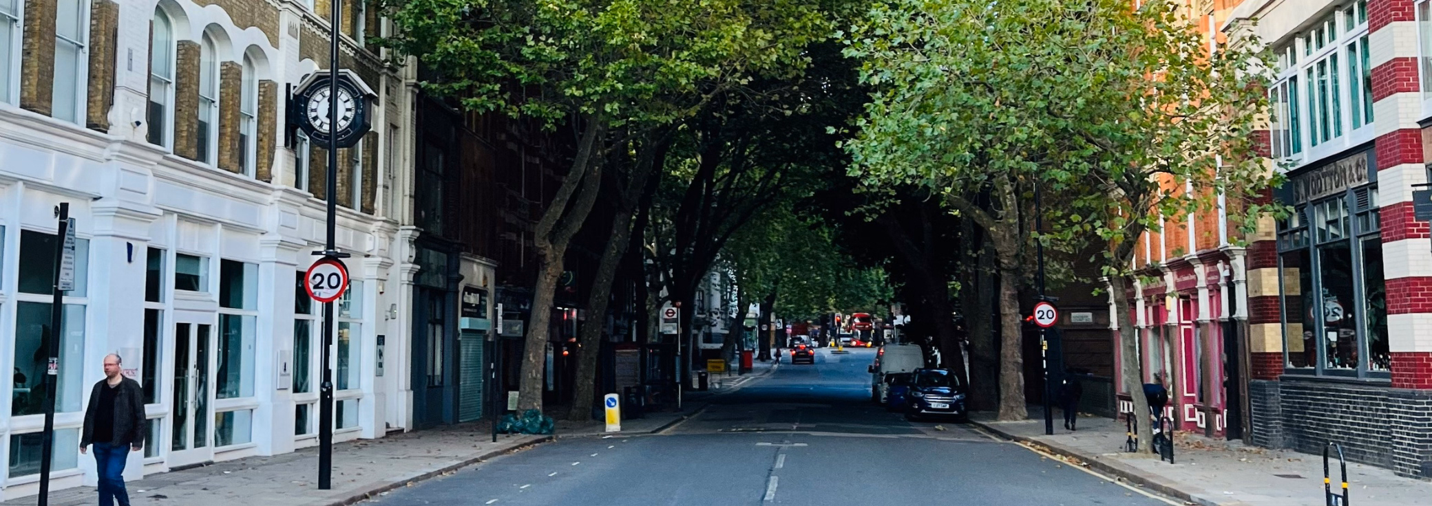 A man walks past white and red brick buildings on a street with trees, cars, a clock, and speed limit signs.