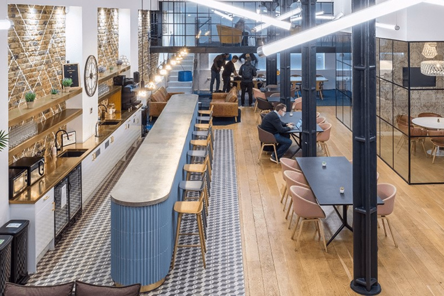 An office kitchen and lounge with people working at black tables with pink chairs, and a blue island counter with wooden stools.