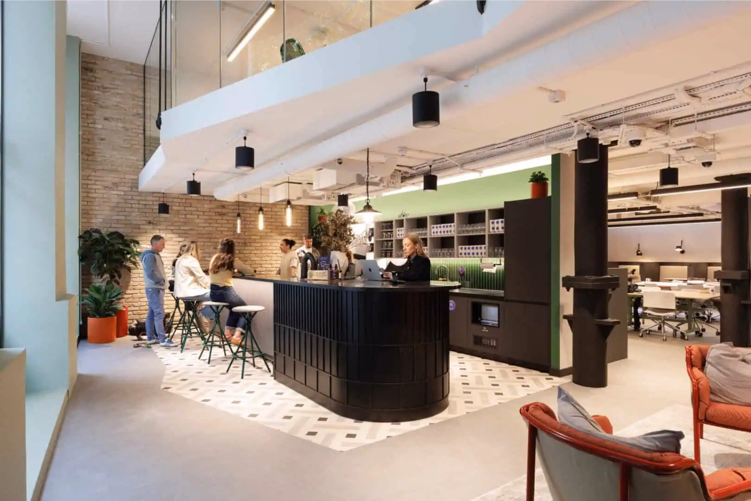 An office kitchen with people talking at a black counter on white stools, and a person using a laptop next to orange armchairs.