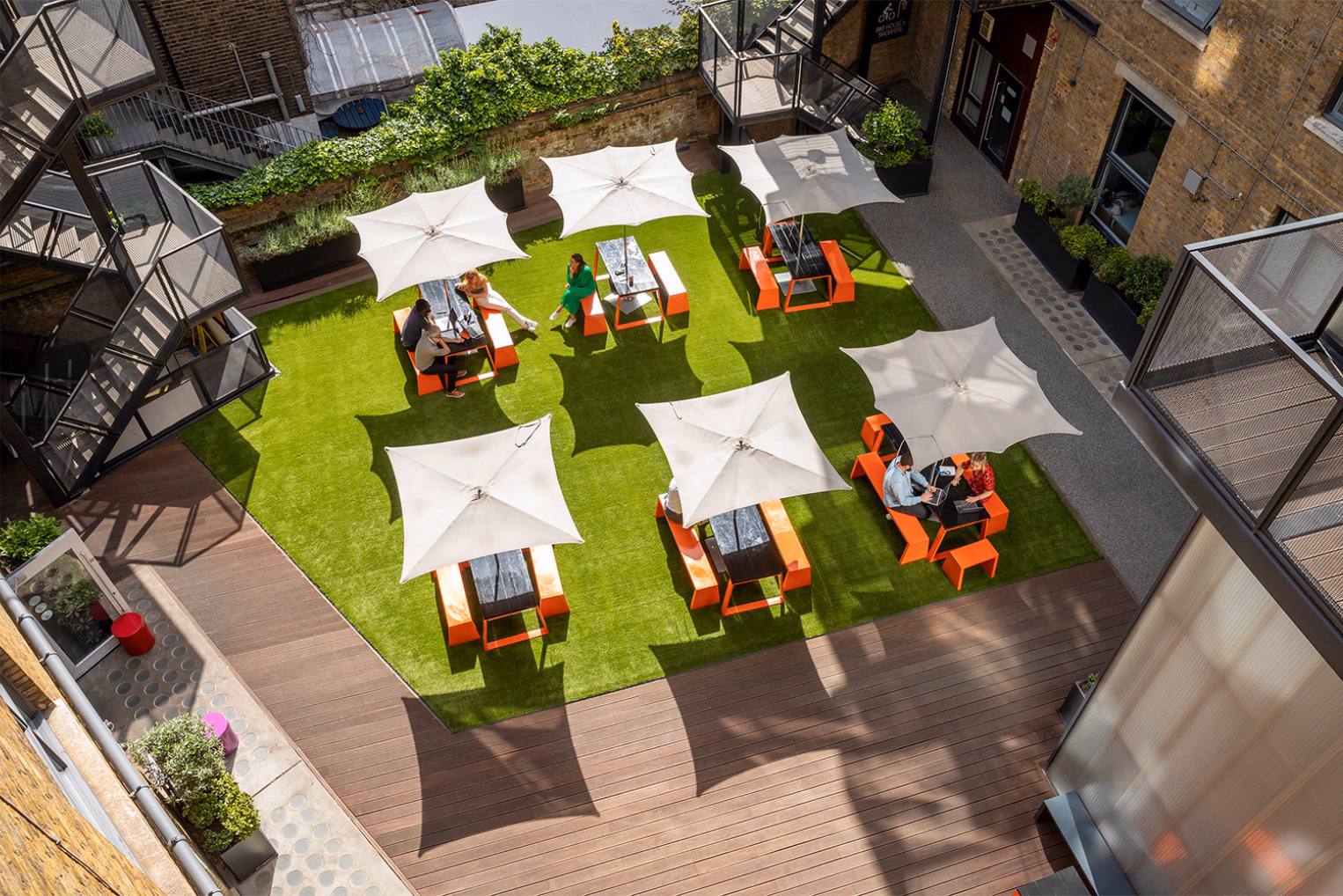 A courtyard in Workspace Clerkenwell Workshops with people sitting at orange benches at black tables with white umbrellas.