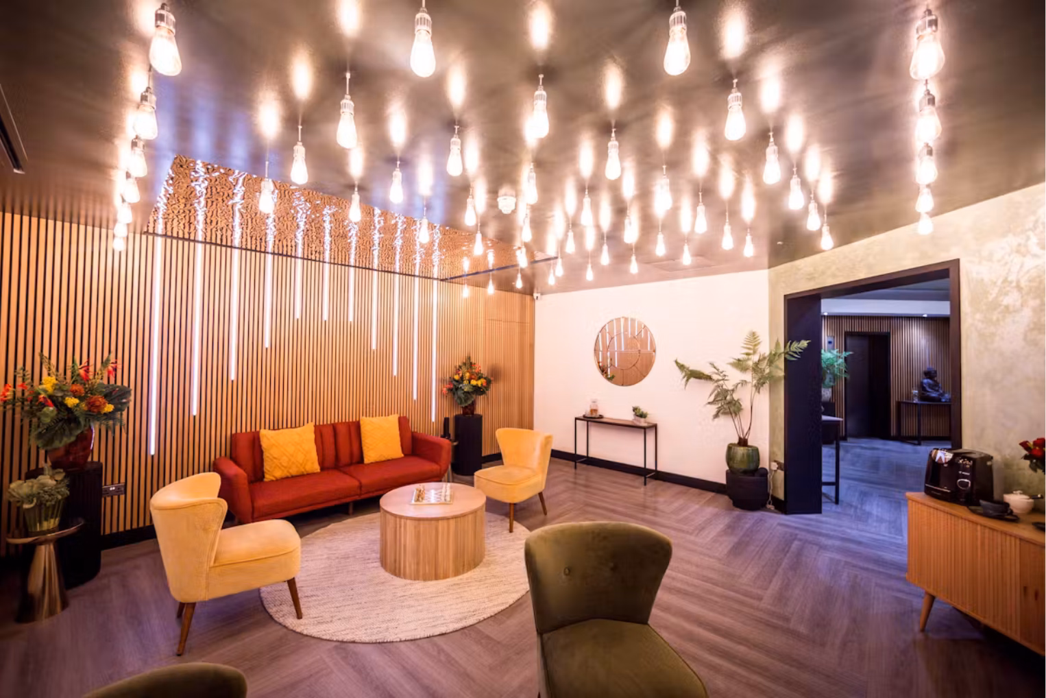 A waiting room with yellow armchairs and a red sofa around a wood coffee table under hanging light bulbs.