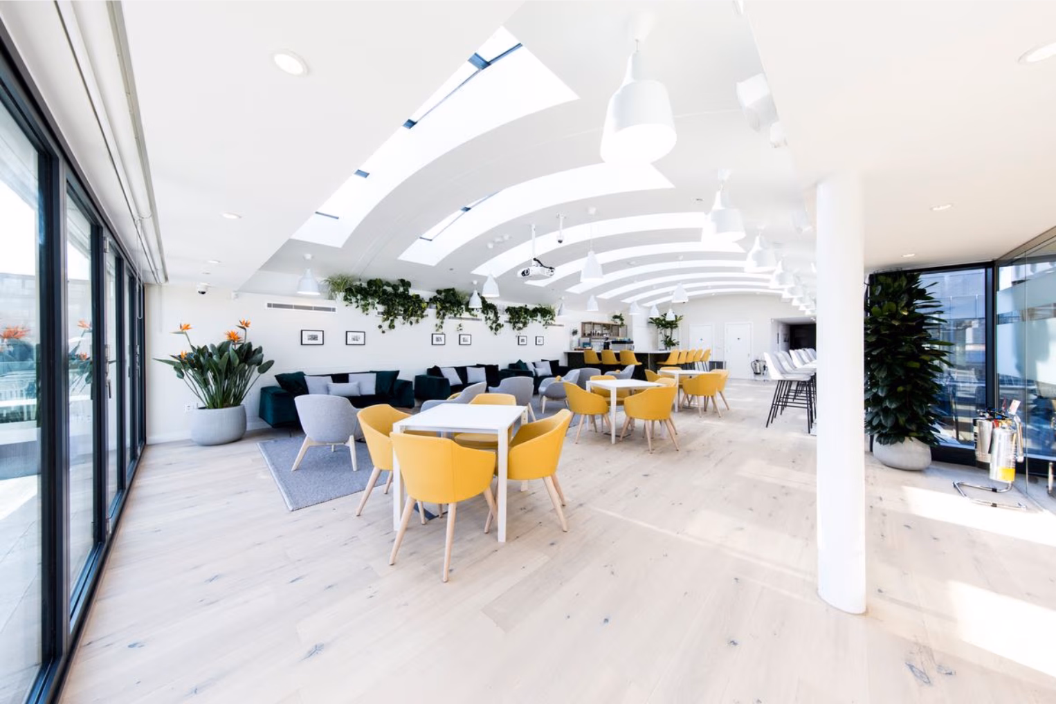 An empty lounge with yellow chairs around white tables, grey armchairs, and green sofas under skylights.