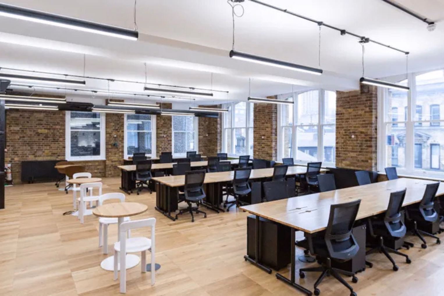 An office space with black chairs at wooden desks, next to white chairs around small round tables.