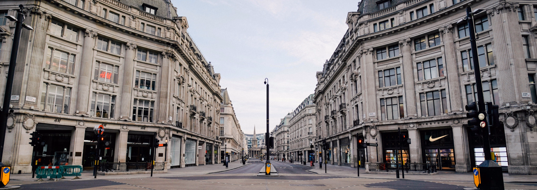 An empty road intersection between stone buildings with an H&M storefront and a Nike storefront under a grey sky.