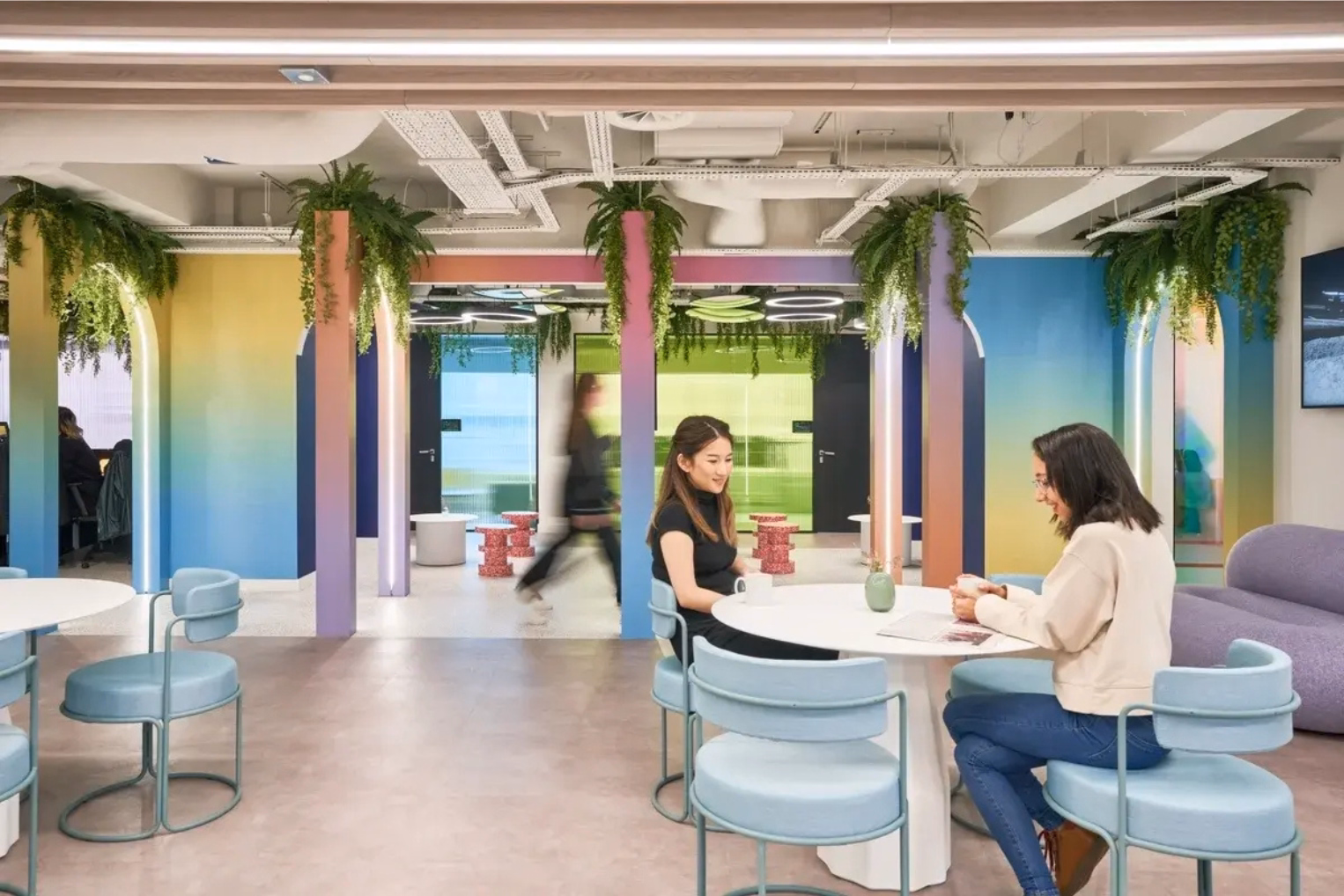 People talk and sit at white tables with light blue chairs near a purple sofa.