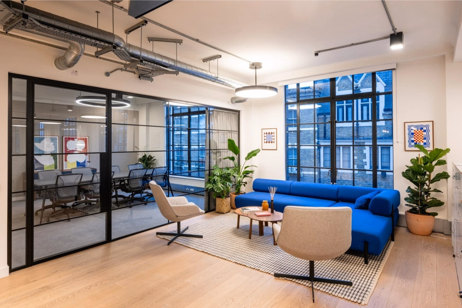 A blue sofa and tan armchairs sit near a brown coffee table, with grey office chairs around a table visible through a glass wall.