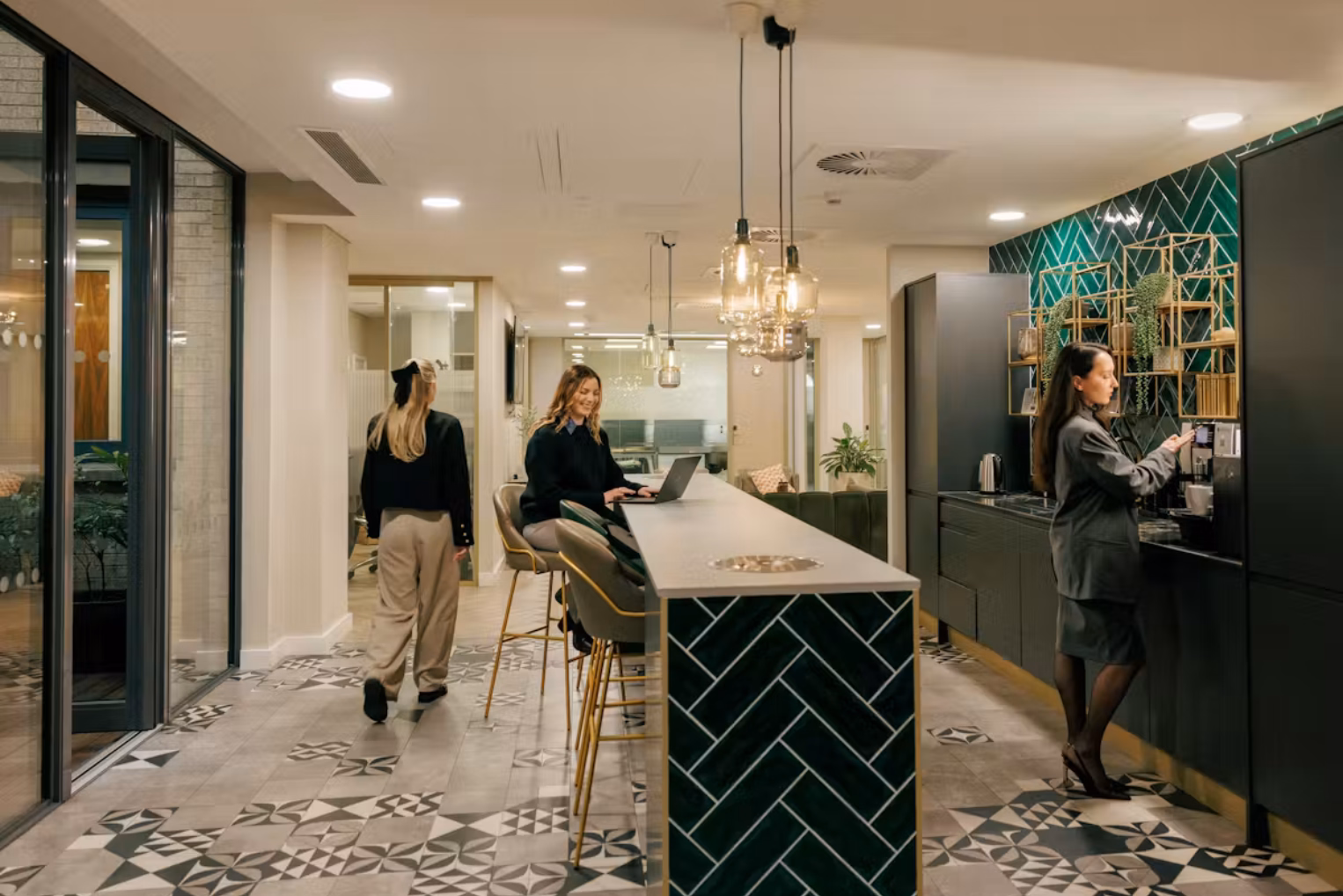 People talk, walk, and use a laptop near a white and green counter with grey stools next in a black and green kitchen.