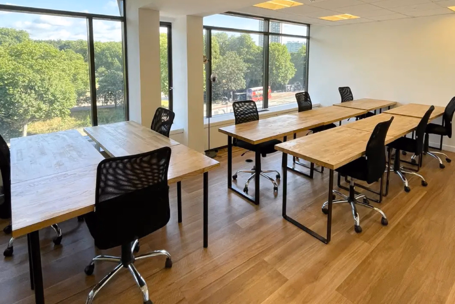 Black office chairs sit at light brown wooden desks in a room with windows overlooking a large amount of green space.