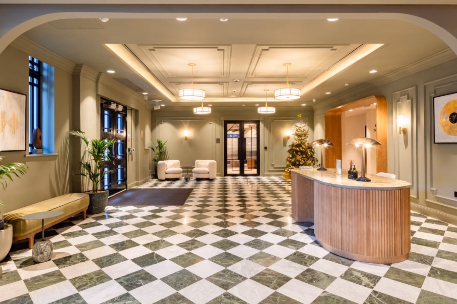 A brown wooden reception desk stands on a checkered floor near white armchairs and an olive coloured bench.