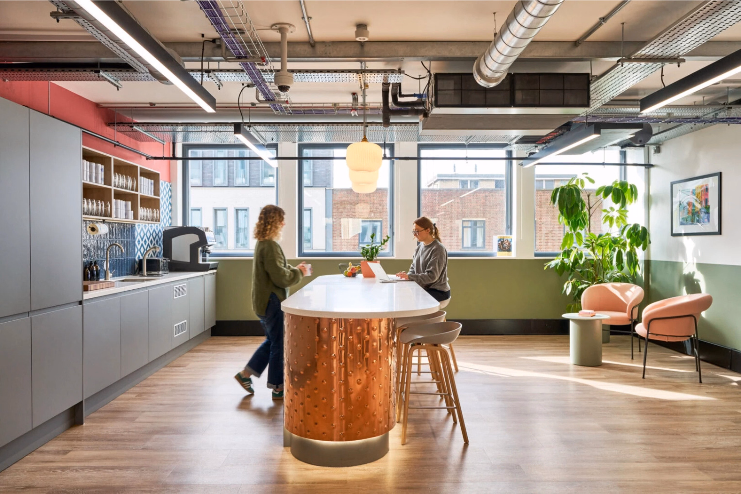 Two women talk and use a laptop at a white and copper counter with grey stools near pink armchairs and a grey kitchen.
