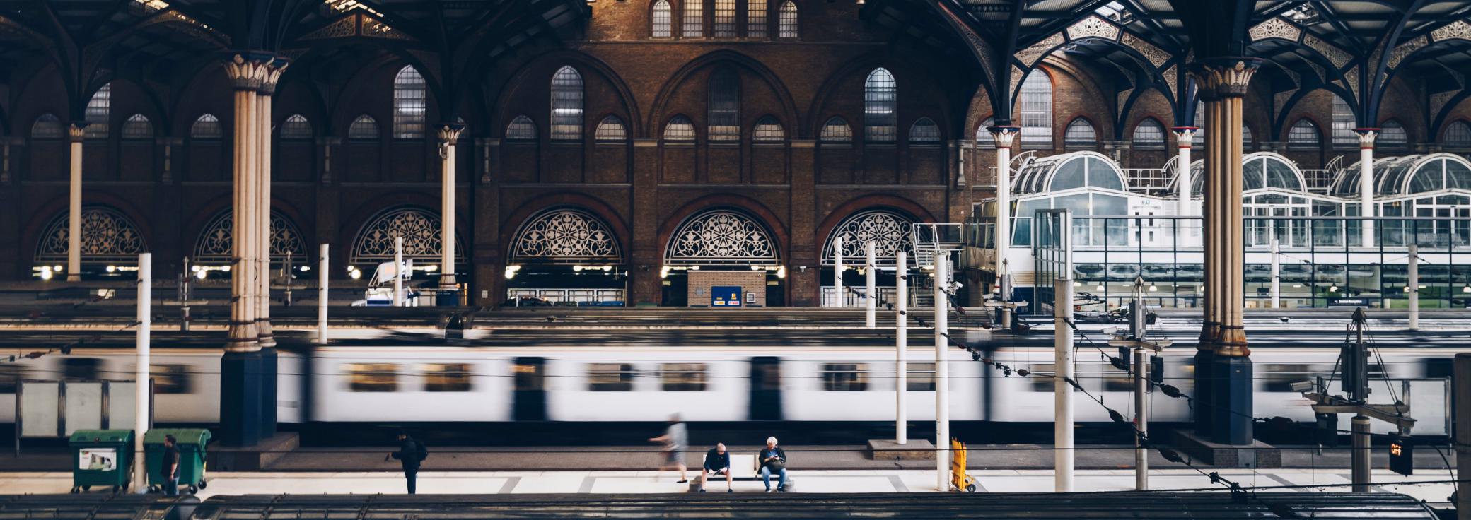 People sit, stand, and walk on a train platform next to a white bench as a train moves past inside a station with brick walls.