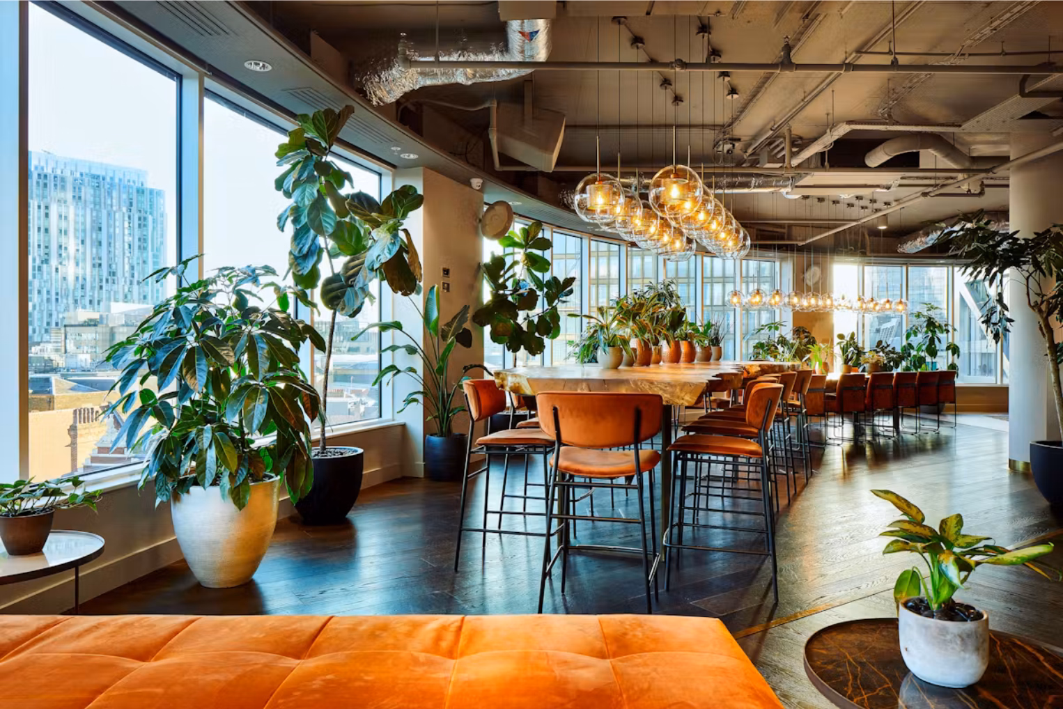 An orange bench and brown stools sit at a long wooden table surrounded by plants near windows overlooking buildings.