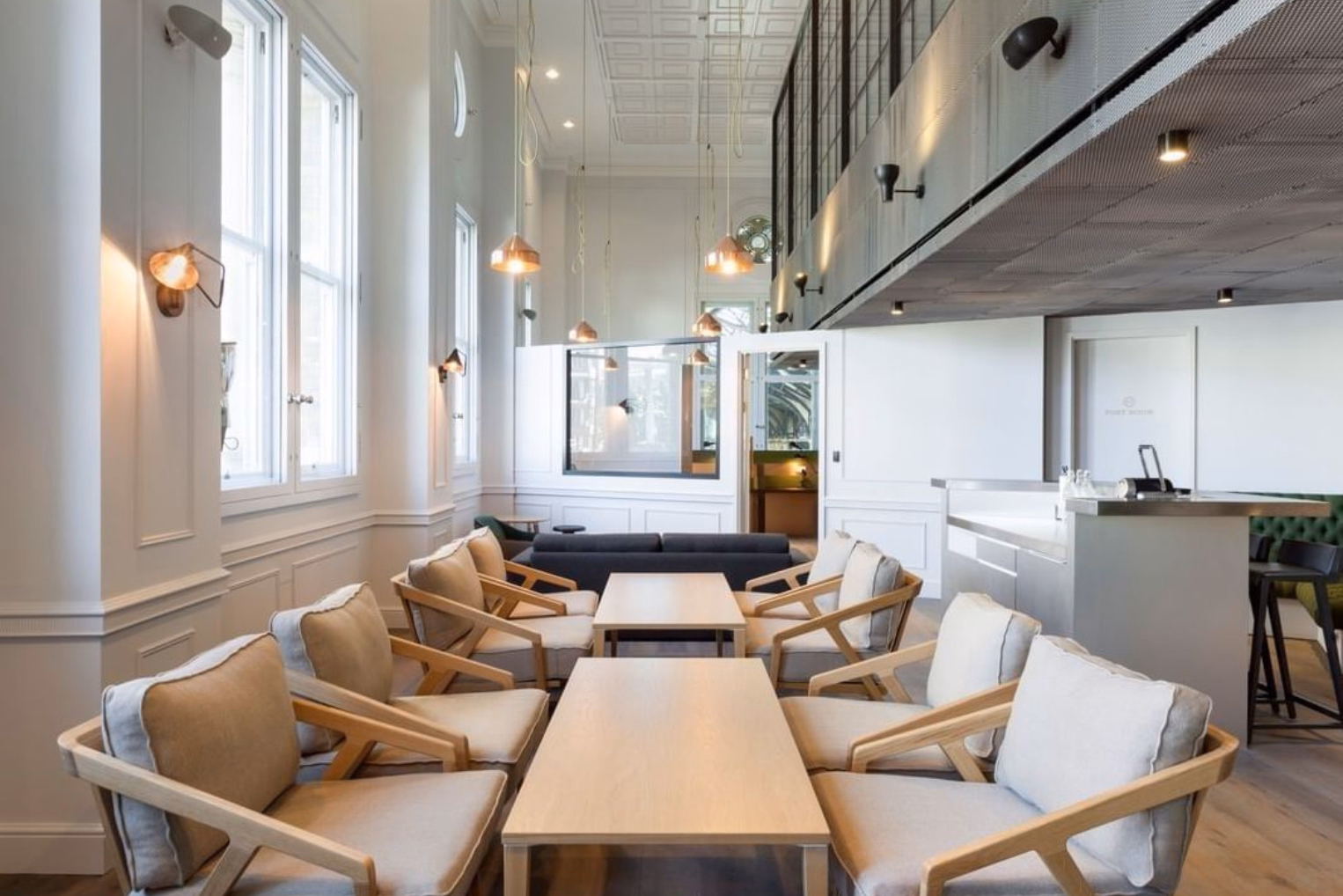 Tan wooden armchairs sit at light brown wooden tables near a black sofa and a grey counter with black stools.