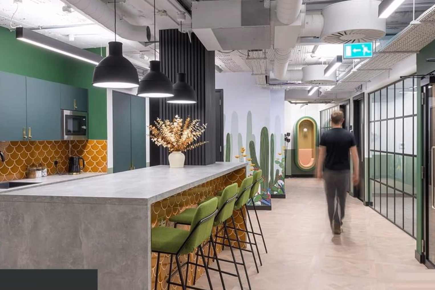 A man walks past a grey marble kitchen counter with green stools near a teal kitchen and a cactus mural.