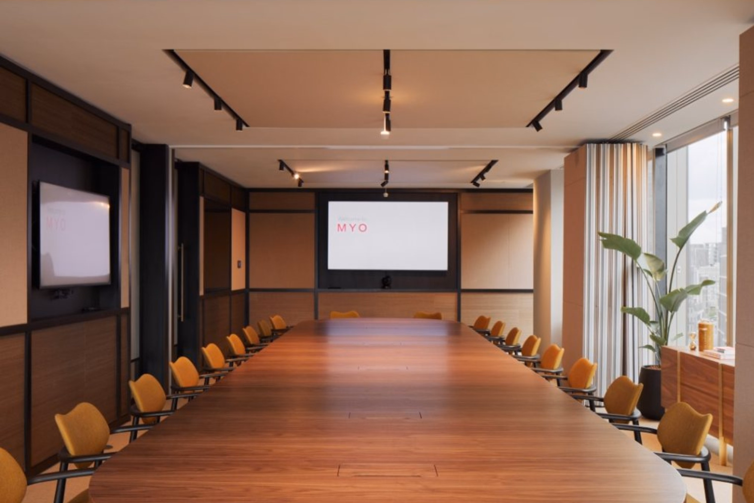 Yellow office chairs sit around a long brown wooden conference table near a screen displaying "MYO" text.