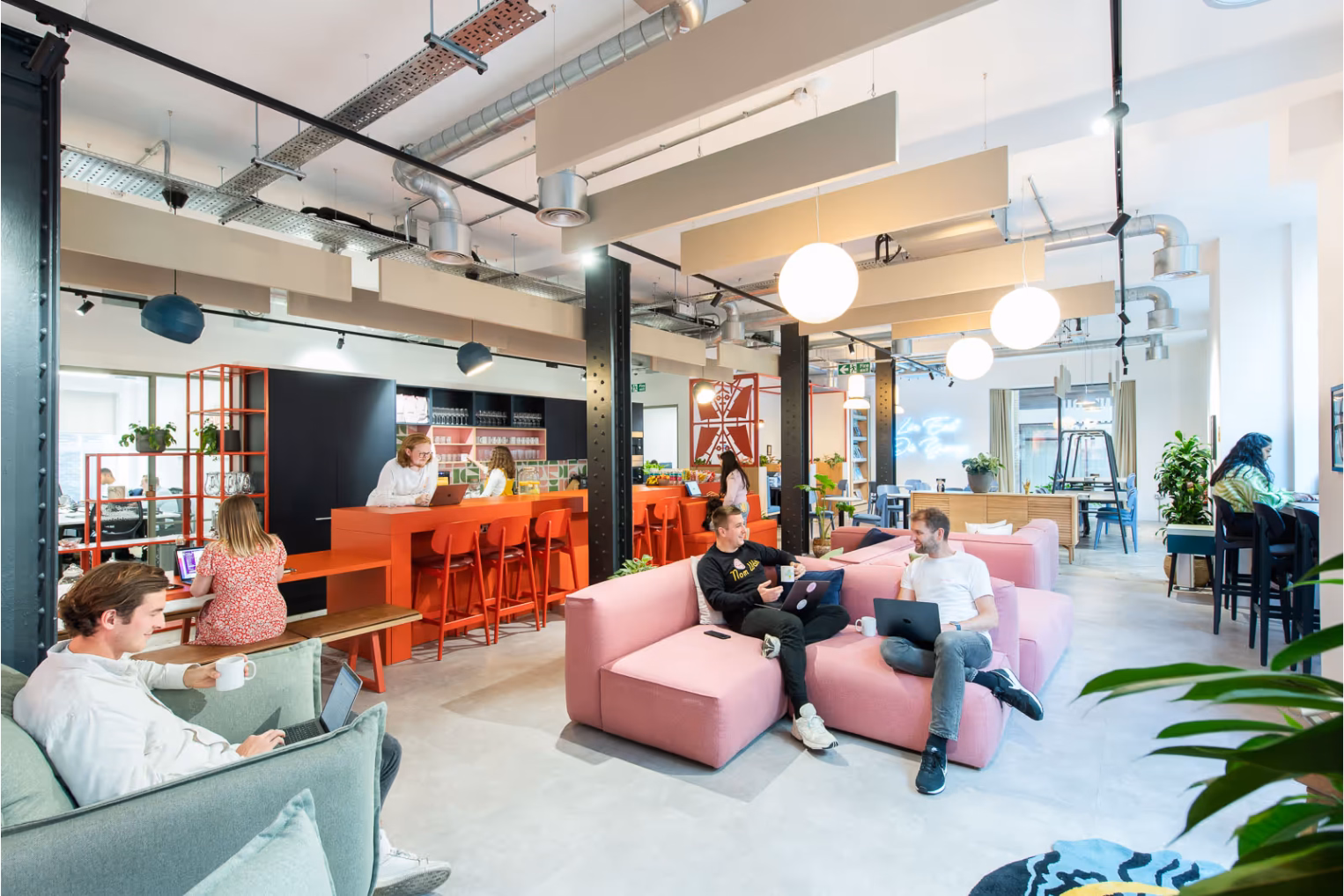 People sit, talk, and use laptops on pink and green sofas near an orange counter with orange stools.