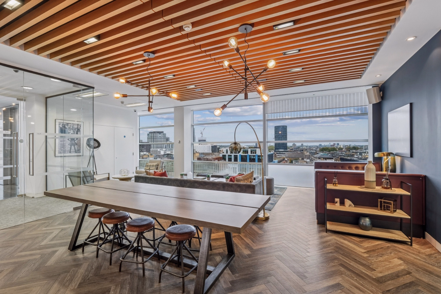 Brown stools sit at a long brown wooden table near a grey sofa and a purple booth by windows overlooking a city skyline.