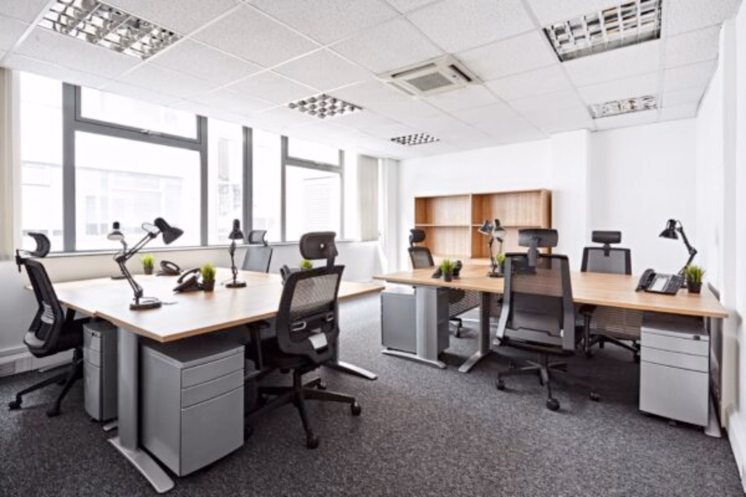 Black office chairs sit at light brown wooden desks with grey filing cabinets in an office room with large windows.