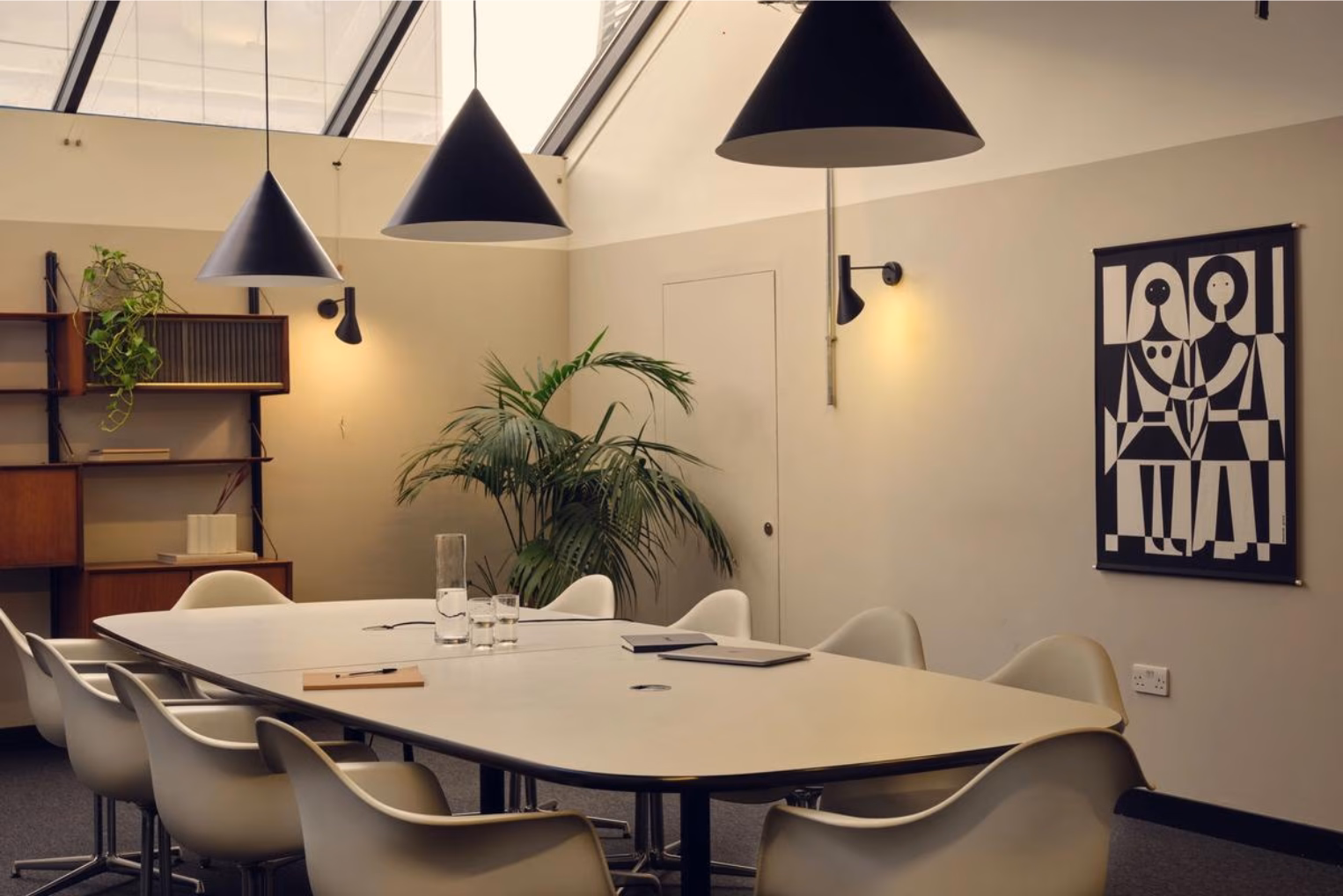 White chairs surround a large white conference table under black cone pendant lights in a meeting room with a skylight.